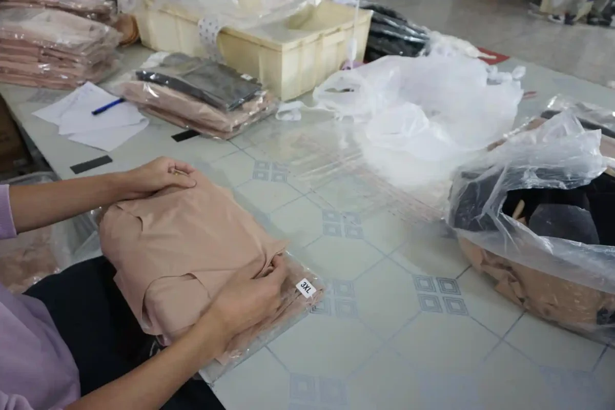 A person packing beige underwear into plastic bags on a factory table surrounded by other packaged items and materials representing the work of underwear suppliers and vendors in the final production stages