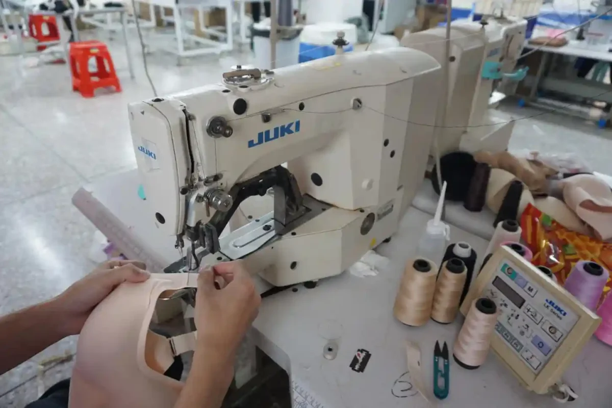 A person operating a Juki sewing machine to stitch a beige bra with various thread spools and sewing tools around highlighting the work of underwear suppliers and vendors in manufacturing